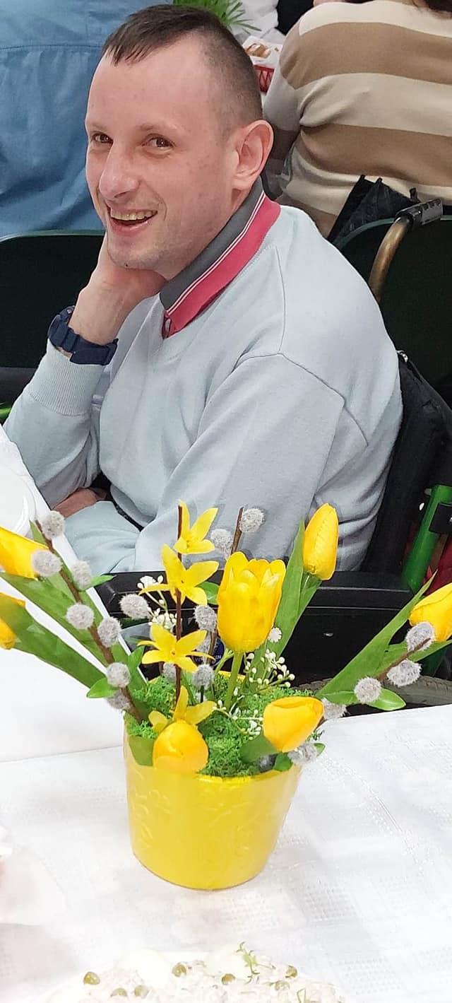 Mężczyzna siedzi na wózku inwalidzkim, uśmiecha się, przed nim na stole stoi bukiet tulipanów.